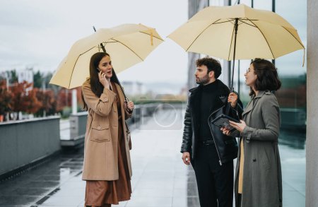 Drei stylische Business-Profis mit gelben Regenschirmen diskutieren an einem regnerischen Tag über die Arbeit im Freien.