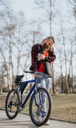 Bearded businessman in casual attire working remotely while standing with his bicycle in an urban park, checking his smart watch.