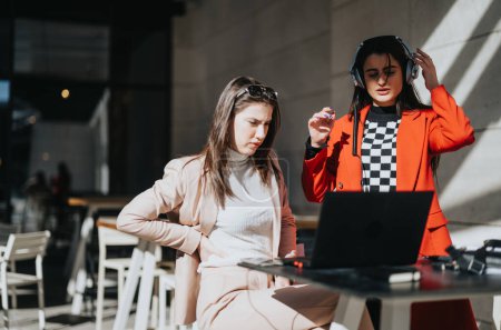 A focused young businesswoman in headphones looks at a laptop while her colleague reads documents at an outdoor cafe table.