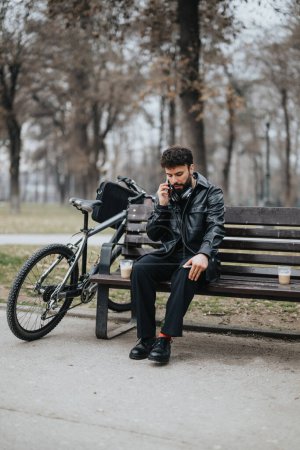 A stylish man in business attire sits on a park bench, working on his smart phone with a bicycle beside him, exuding urban professionalism in a natural setting.