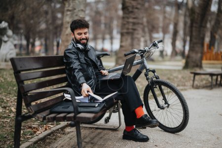 Stylish male entrepreneur multitasking remotely with a laptop and notepad in a city park, accompanied by his bike.