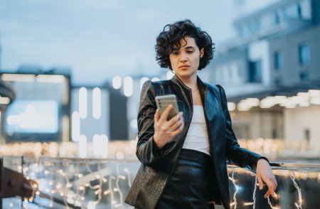 Stylish young woman using smart phone in urban evening scene with festive lights.