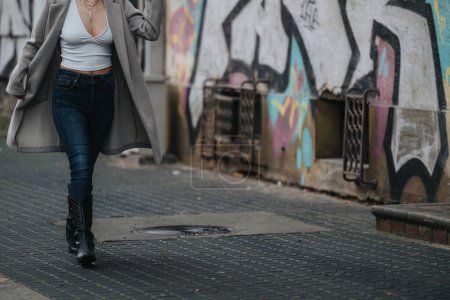 Mujer joven de moda paseando con confianza en un camino empedrado. Estilo urbano con abrigo gris y jeans, fondo de graffiti que transmite elegancia casual.