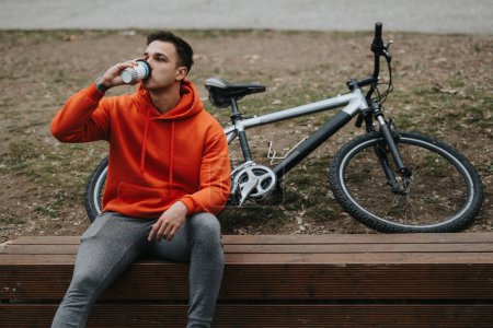 Active young adult resting on a bench in a park, drinking from a cup, with a mountain bike beside him, on an overcast day