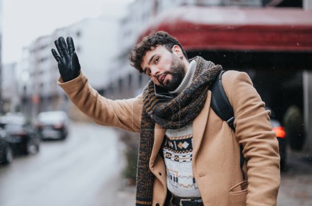 Elegant gekleideter junger Mann winkt an einem winterlichen Tag nach einem Taxi. Hes draußen, zeigt ein Gefühl der Dringlichkeit und Entschlossenheit inmitten von Schneeflocken.