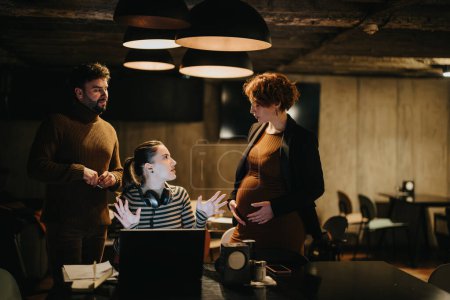 Stunning, short haired woman talking about her pregnancy with her colleagues while working late at night in modern office setting.