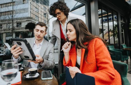 Drei Geschäftsleute diskutieren in einem Outdoor-Café über ein Tablet, auf dem Tisch liegen Kaffee und Arbeitsgrundlagen.