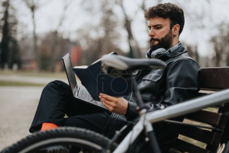 Konzentrierter Geschäftsmann in lässiger Kleidung, vertieft in das Lesen von Dokumenten, während er draußen auf einer Bank sitzt, neben ihm ein Laptop.