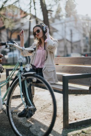 A fashionable young woman in sunglasses listens to music with headphones, sitting on a park bench with her bike.