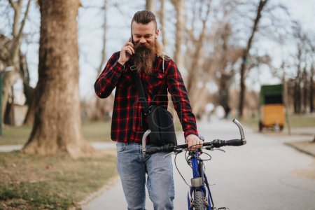 A bearded businessman in casual attire is on a phone call, holding his bicycle in a green urban park setting, portraying a modern remote working lifestyle.