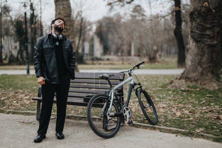 Elegante hombre de negocios en traje negro de pie junto a un banco del parque con una bicicleta, teniendo en el entorno tranquilo.
