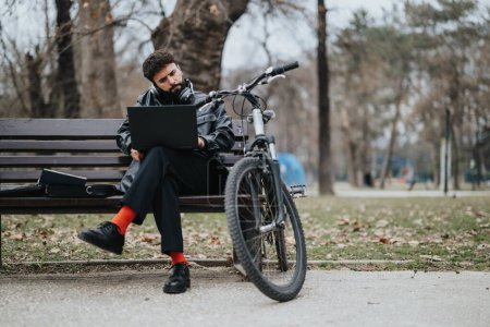 Stilvoller männlicher Profi mit Laptop und Fahrrad übernimmt ferngesteuerte Arbeit inmitten der Ruhe eines Stadtparks.