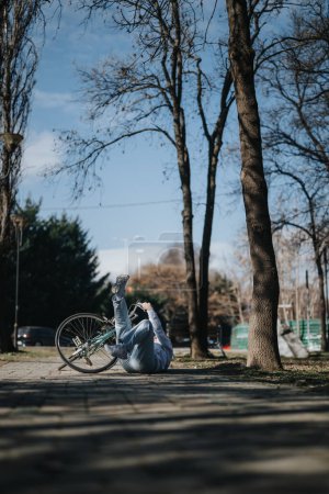 Foto, das einen unerwarteten Moment festhält, als eine Frau in einem ruhigen Park, umgeben von kahlen Bäumen und einem klaren Himmel, von ihrem Fahrrad fällt.