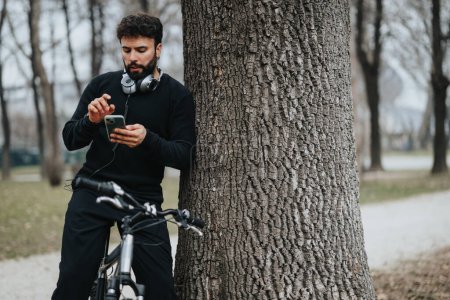 Ein bärtiger Mann lehnt an einem Baum, während er sein Handy kontrolliert und sich mit seinem Fahrrad im Park ausruht. Er trägt lässige Sportbekleidung und Kopfhörer um den Hals.