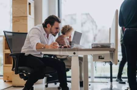 Ein fleißiger Mann in weißem Hemd vertieft in seine Arbeit an einem Laptop in einem modernen Büroraum mit Kollegen im Hintergrund.