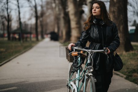 A professional woman in a leather jacket stands with her bike in a city park, reflecting an eco-friendly commute.