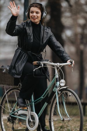 Stylish and confident young businesswoman wearing a black outfit and headphones, waving while riding a bicycle in an urban setting.