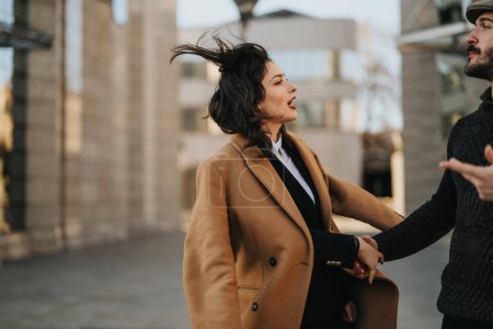 A professional woman in a camel coat laughs during a dynamic discussion with a male coworker on a city street, conveying teamwork and engagement.