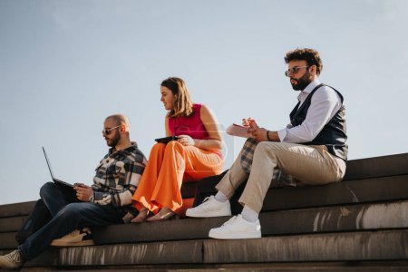 Drei junge Berufstätige, die mit Laptops und Tablets im Freien auf einer Treppe sitzen und ein Geschäftsgespräch führen.