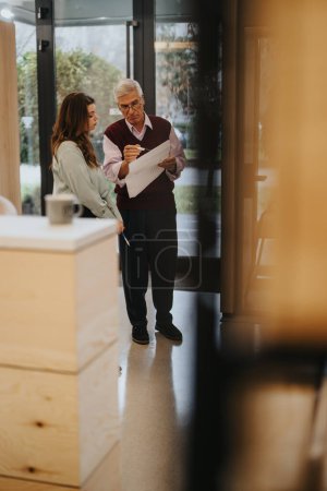 Ein erfahrener älterer Mann begutachtet ein Dokument mit einer fokussierten jungen Mitarbeiterin in einem hellen, modernen Büroumfeld, das Mentorenschaft und Zusammenarbeit symbolisiert.