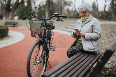 Reife Rentnerin genießt Freizeit im Park mit Fahrrad, Smartphone im Sitzen auf Bank