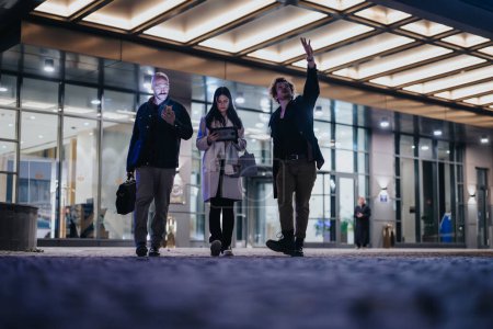 Entrepreneurs exiting an office building, engaged in an informal discussion with one person gesturing upwards, in an urban night setting.