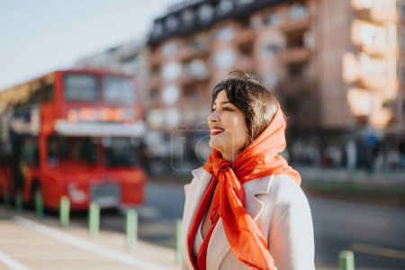 Smiling woman with red scarf on city street, red double-decker bus in background.