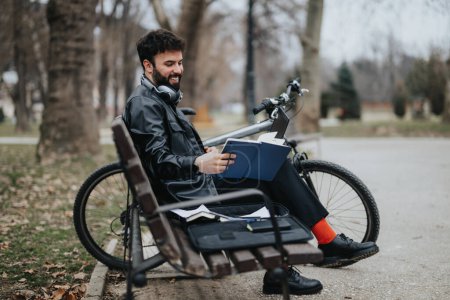 Stylish businessman enjoys working remotely from the park, using a phone and notebook with his bicycle nearby.