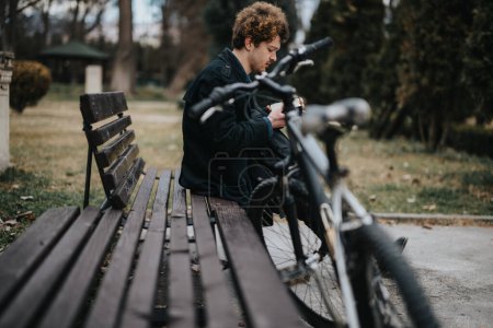 Un jeune professionnel ou entrepreneur travaillant à distance sur un téléphone intelligent, à côté d'un vélo sur un banc de parc tranquille.