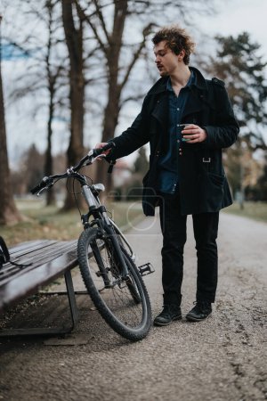 A young, professional man stands by his bike in a park, taking a break from the busy workday.