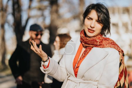 Outdoor portrait of a stylish young woman with a vibrant scarf enjoying a sunny day in the park, casually conversing with friends in the background.