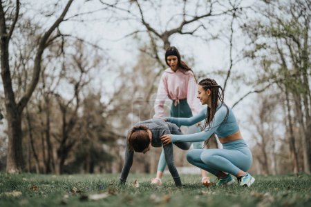 A professional fitness instructor guides two women during a stretching exercise routine in a serene park setting.