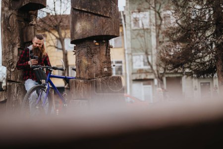 Un homme barbu en chemise à carreaux s'arrête pour prendre un café et utiliser son téléphone intelligent à vélo dans un cadre urbain.