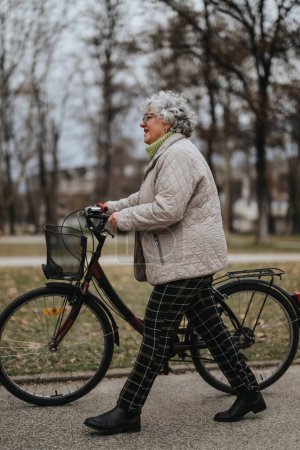 Aktive Rentnerin mit Brille und einem Lächeln spaziert mit dem Fahrrad durch eine ruhige Parklandschaft.