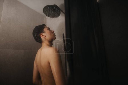 Rear view of a young adult male standing under a running shower head in a contemporary bathroom setting, depicting hygiene and daily routine.