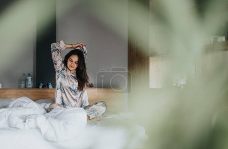 Eine entspannte junge Frau dehnt sich, während sie auf einem Bett sitzt und vermittelt ein Gefühl von Frieden und Komfort in einer häuslichen Umgebung.