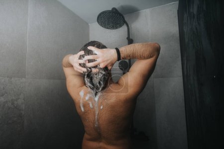 Die Rückansicht eines Mannes, der sich unter einer laufenden Dusche mit Shampoo den Kopf schrubbt, stellt Körperpflege und Sauberkeit dar.