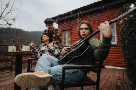 Young adults relax with guitars and coffee on a wooden porch by a rustic cabin, sharing a moment of music and laughter.