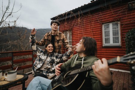 Eine Gruppe von Freunden versammeln sich auf einer Holzveranda, spielen Gitarre und genießen den Blick auf die Berge von ihrem rustikalen Hüttenaufenthalt.