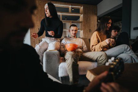 A group of youthful friends gathered around a bowl of snacks, socializing and playing guitar in a cozy living room setting.