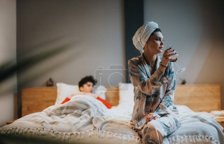 Komfort zu Hause mit einer Frau, die sich in kuscheligen Pyjamas entspannen und ein Glas Wein in einer ruhigen Schlafzimmeratmosphäre genießen kann.