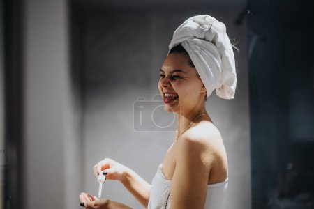A cheerful young woman wrapped in a towel, with another on her head, is singing and having fun while holding a dropper bottle in a modern bathroom setting.