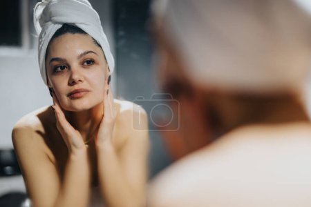 Serene female in her 30s performing skincare routine, observing her complexion in the mirror with a thoughtful expression.