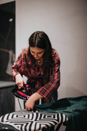 Focused woman ironing a green shirt on a stylish ironing board with a geometric cover in a household setting.