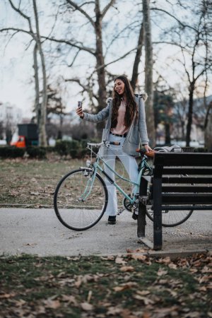 A cheerful businesswoman in casual attire with her bike captures a selfie moment in a serene park.