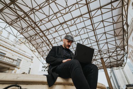 Empresario enfocado que trabaja en un ordenador portátil mientras está sentado al aire libre, con arquitectura moderna y una cubierta geométrica.