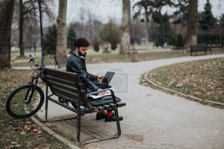 Konzentrierter männlicher Unternehmer mit Laptop auf einer Parkbank neben seinem Fahrrad, der mobile Arbeit und Flexibilität verkörpert.