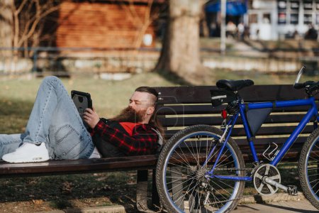 Un homme d'affaires barbu en tenue décontractée aime travailler à distance dans un parc urbain, en utilisant une tablette avec son vélo à proximité.