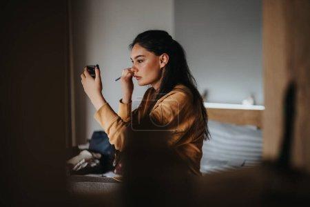 An indoor shot capturing a relaxed young woman engrossed putting make up while using her phone as a mirror.