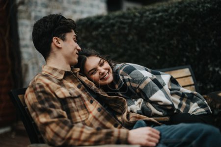 Young couple in love enjoying a warm embrace outdoors, snuggled under a checkered blanket on a bench, exuding happiness and connection.